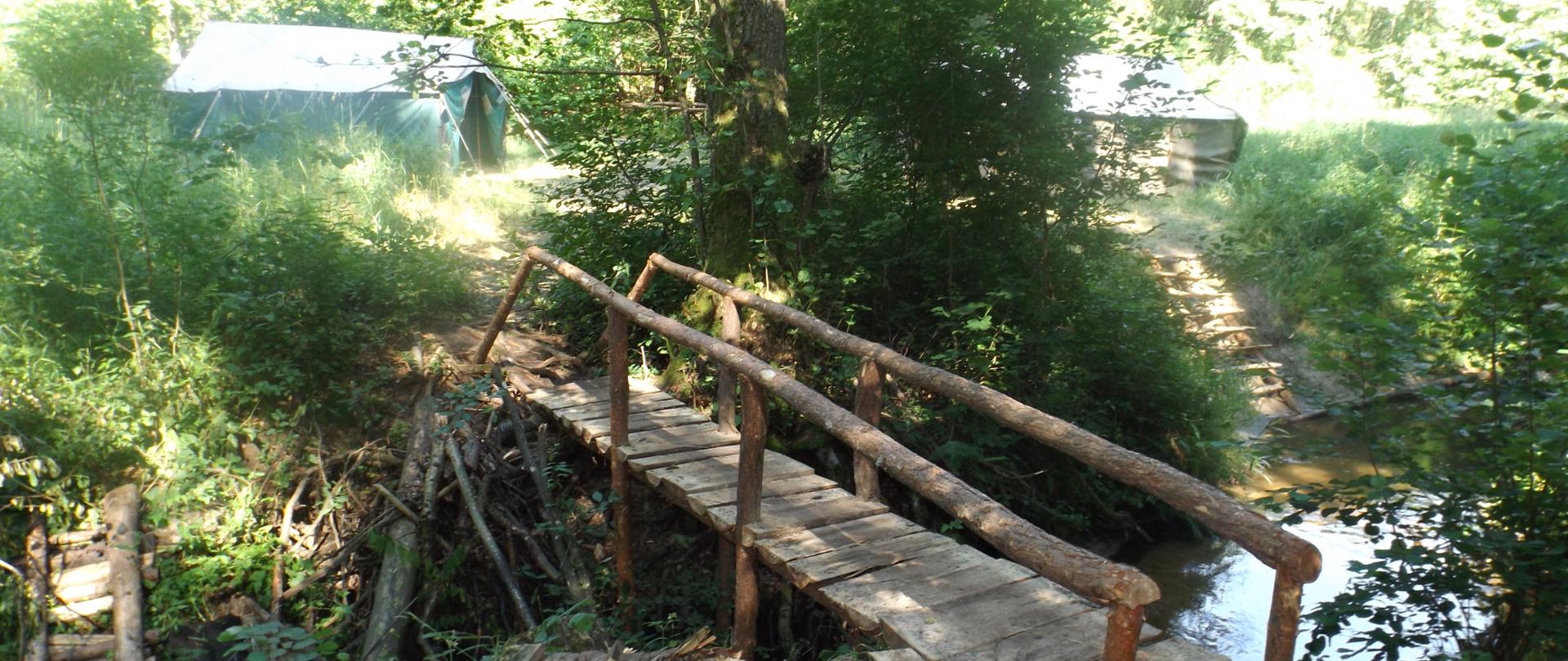 Zdjęcie przedstawia mostek prowadzący do obozu harcerskiego. Obok widać połamane drzewa i krzaki.