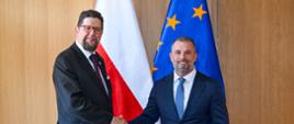 Wiceminister Szeptycki stoi obok mężczyzny w garniturze, podają sobie ręce, za nimi przed wyłożoną drewnem ścianą flagi Polski i UE.