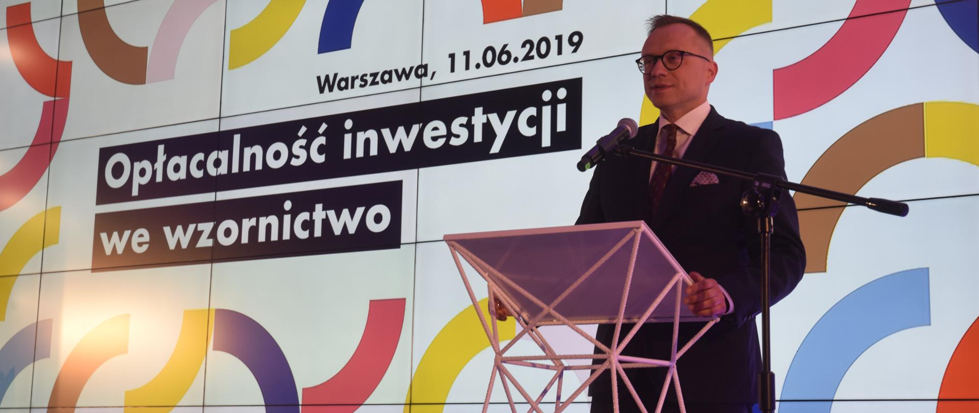 Podczas konferencji wiceminister Artur Soboń stoi na scenie w mównicy przed mikrofonem. Za nim ekran z wyświetlonym napisem „Opłacalność inwestycji we wzornictwo, Warszawa, 11.06.2019"