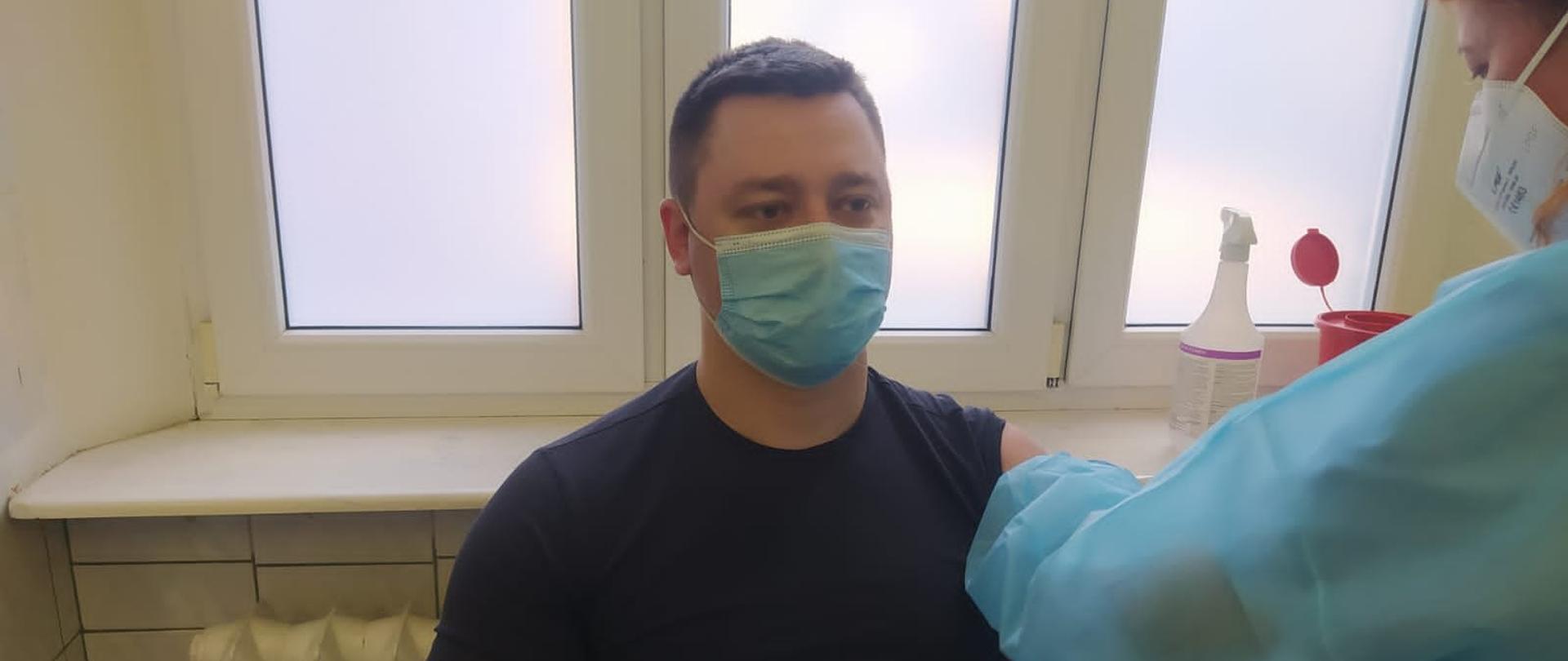 Strażak w czarnej koszulce i maseczce na twarzy siedzi w gabinecie a pielęgniarka w niebieskim stroju i maseczce na twarzy wykonuje szczepienie.