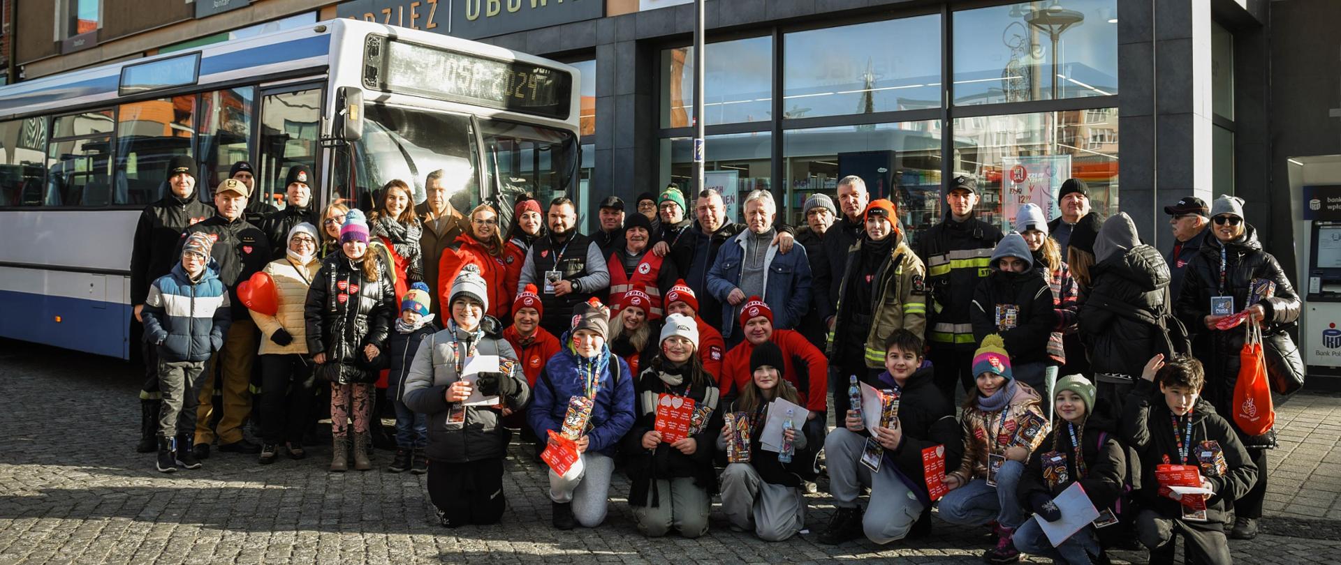 Czterej funkcjonariusze Komendy Powiatowej Państwowej Straży Pożarnej w Lęborku na zdjęciu grupowym wraz z innymi osobami, w tym wolontariuszami zaangażowanymi w przebieg 32. Finału Wielkiej Orkiestry Świątecznej Pomocy na Placu Pokoju w Lęborku. W tle biało - niebieski autobus z widocznym napisem WOŚP 2024 oraz witryny sklepów.
