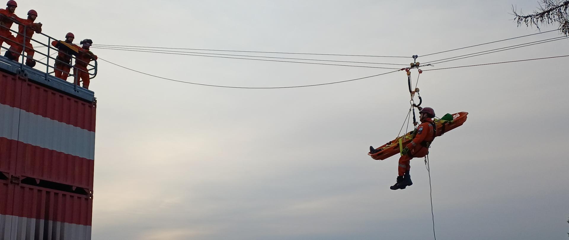Zdjęcie przedstawia ćwiczenia służb ratunkowych na otwartej przestrzeni w pochmurny dzień. Na pierwszym planie, po lewej stronie, widać wysoką konstrukcję z czerwono-białych kontenerów transportowych. Na jej szczycie, zabezpieczonym barierką, stoi grupa czterech ratowników w pomarańczowych kombinezonach i kaskach.
Od szczytu kontenerów w prawą stronę biegną liny nośne, na których zawieszony jest ratownik transportujący nosze kubełkowe. Ratownik znajduje się w powietrzu, podpięty do systemu lin; siedzi w uprzęży tyłem do kontenerów, podtrzymując przed sobą poziomo ułożone nosze. Z noszy zwisa pionowo w dół długa, cienka lina asekuracyjna. W tle widać zamglone niebo z bladą tarczą słońca nisko nad horyzontem, sylwetki bezlistnych drzew oraz dachy niskich budynków przemysłowych.