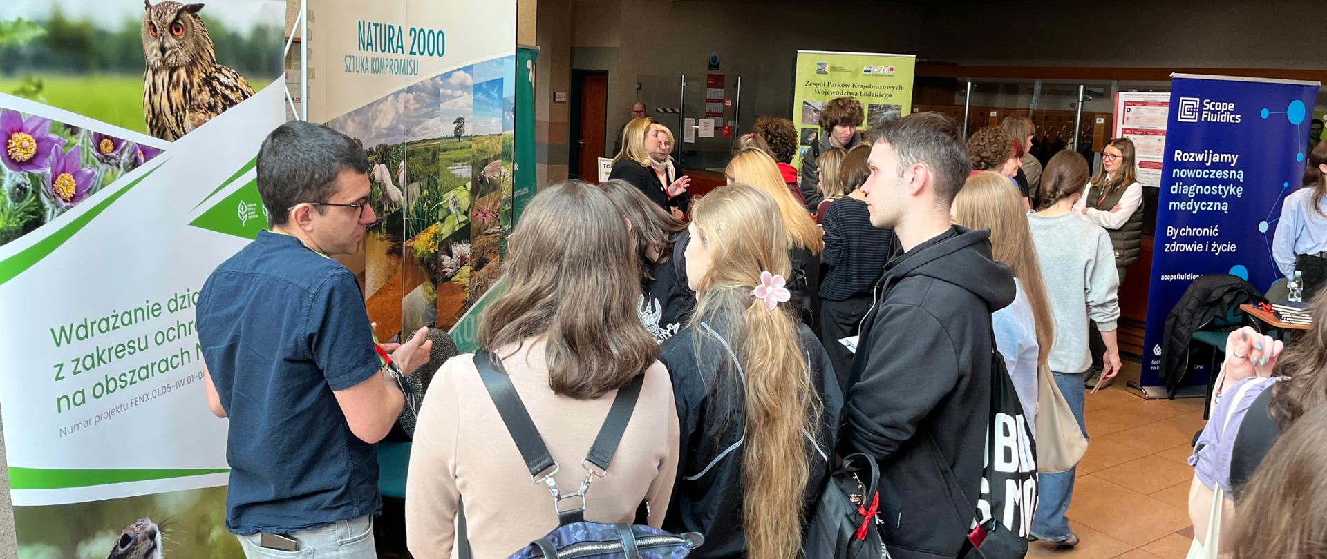 Grupa młodych ludzi zgromadzona przy stoisku informacyjnym dotyczącym ochrony przyrody (Natura 2000) podczas wydarzenia w budynku; uczestnicy rozmawiają z prezentującym, w tle widoczne inne stoiska i banery.