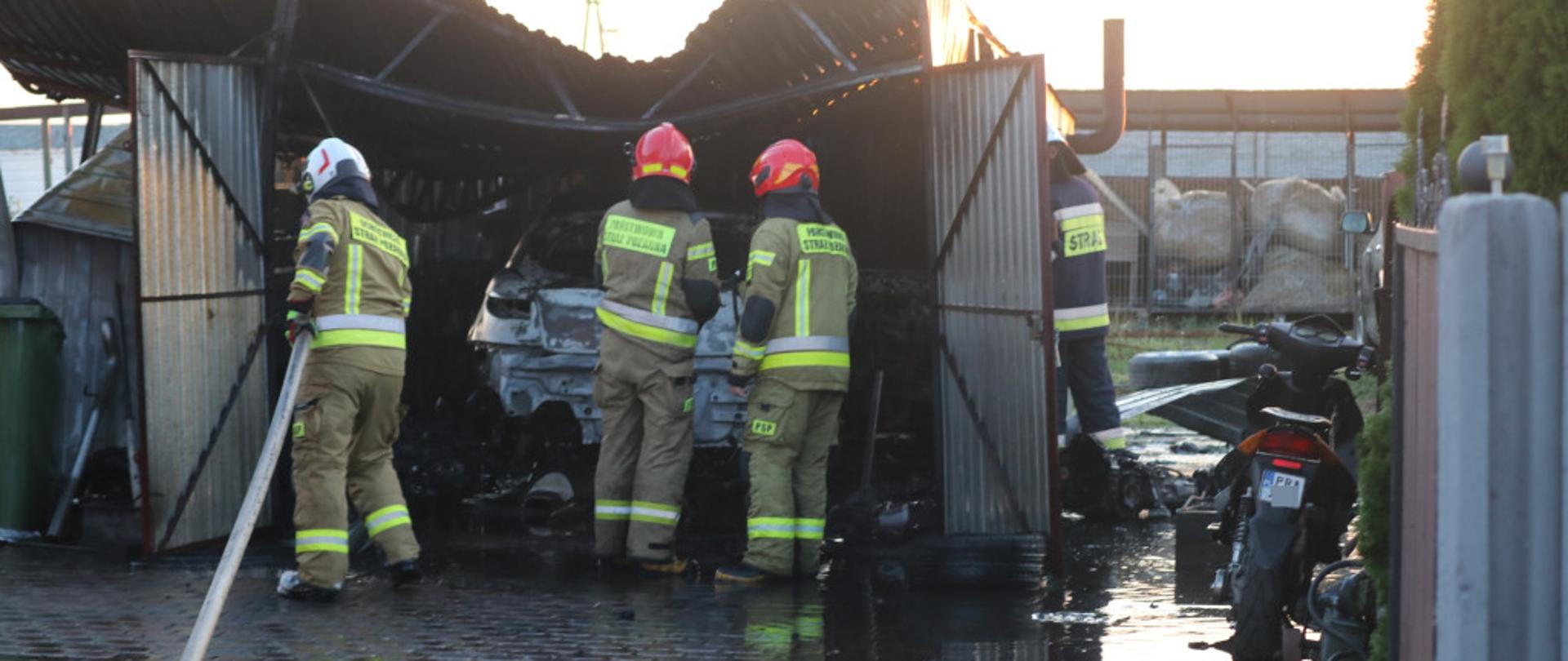Zdjęcie przedstawia strażaków OSP oraz druhów którzy dogaszając wnętrze garażu w którym doszło do pożaru. Jest to garaż blaszany. W garaży znajduje się spalony samochód osobowy. Z prawej strony przed garażem stoi skuter. 