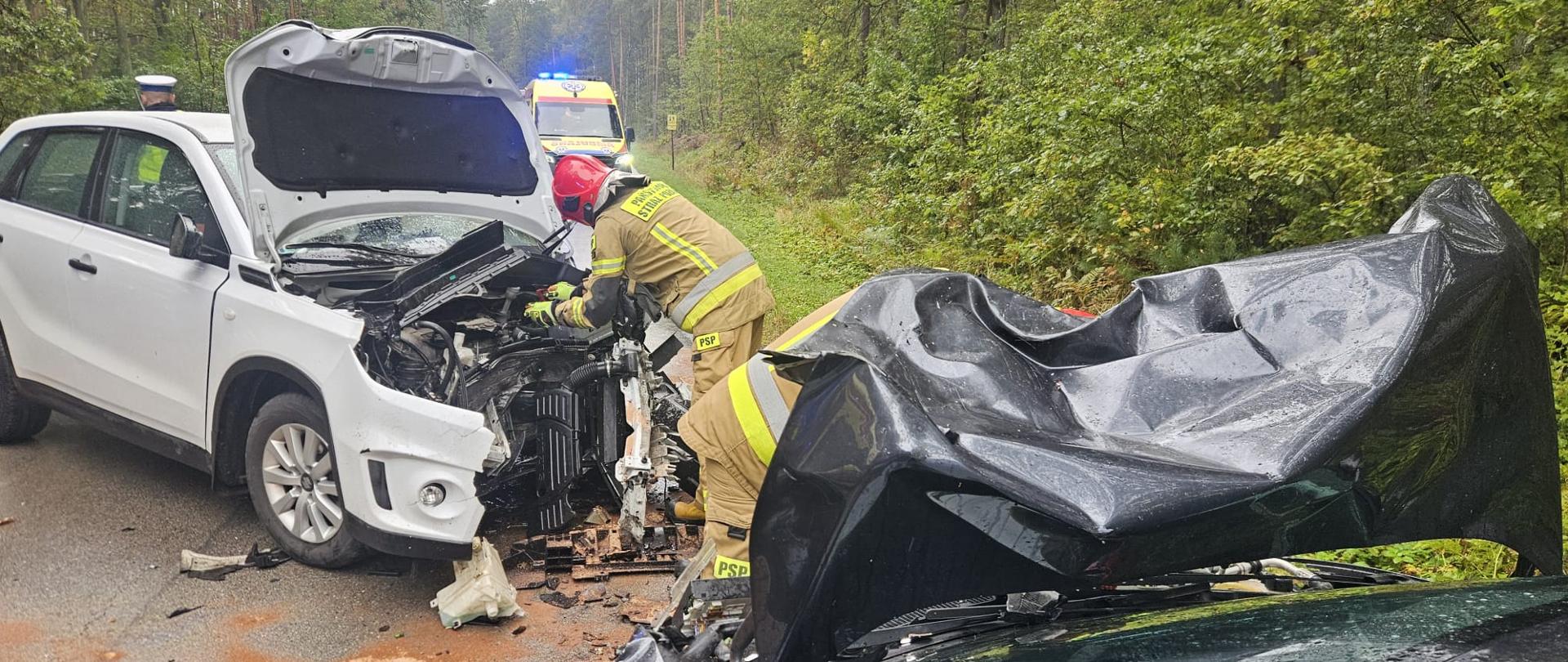 Zdjęcie przedstawia rozbity samochód osobowy w przydrożnym rowie melioracyjnym. Samochód ma podniesioną maskę oraz drzwi kierowcy. W tle widnieje karetka pogotowia.