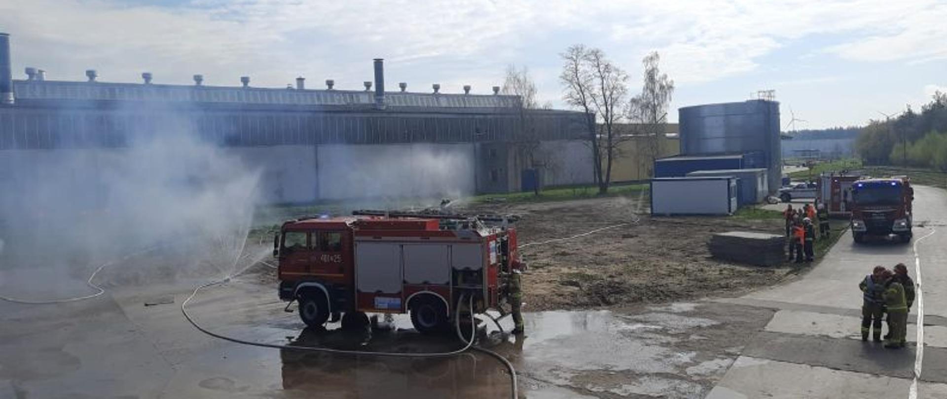 Widok na miejsce prowadzenia działań z drugiej strony. W oddali zakład produkcyjny. Na pierwszym planie dwa wozy strażackie. Do jednej z linki gaśniczych podłączona kurtyna wodna.