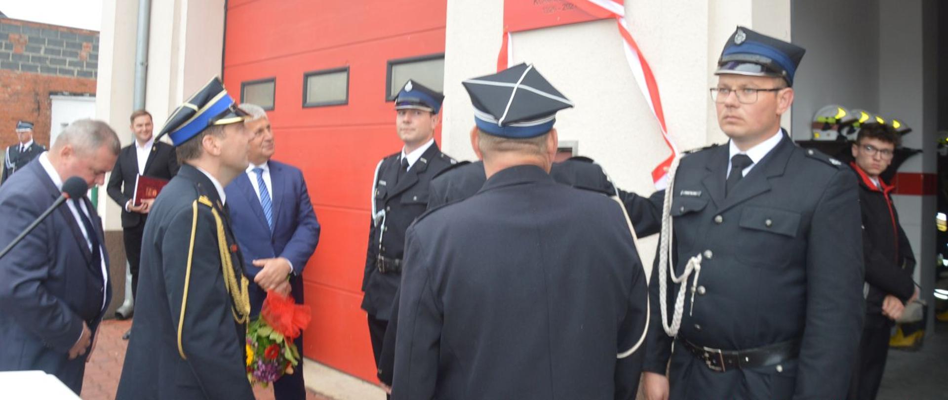 Jubileusz 95-lecia OSP Konary. Plac przed remizą strażacką. Trwa odsłanianie pamiątkowej tablicy, która jest zainstalowana na froncie siedziby OSP. Przy tablicy wartę pełni dwóch druhów, a odsłonięcia dokonują prezes OSP w Konarach i zaproszeni goście.