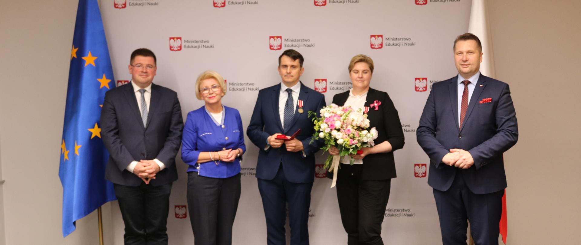 Wspólne zdjęcie Ministra Edukacji i Nauki Przemysława Czarnka, Marzeny Machałek Sekretarza Stanu w MEiN oraz Tomasza Rzymkowskiego Sekretarza Stanu w MEiN z odznaczonymi Katarzyną i Mateuszem Kłoskami. W tle ścianka wierunkowa z logotypami MEiN oraz flagi Polski i Unii Europejskiej