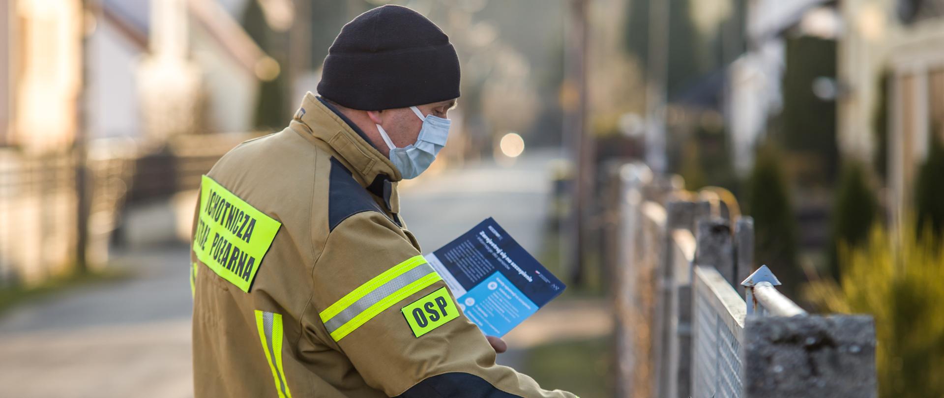 Zdjęcie przedstawia strażaka OSP wrzucającego ulotkę propagujące wiedzę na temat profilaktyki oraz szczepień przeciwko COVID-19. Strażak wrzuca ulotkę prawą ręką do skrzynki na listy. W lewej ręce trzyma pozostałe ulotki. Strażak ubrany w ubranie specjalne koloru piaskowego, czarną czapkę i jasną maseczkę na twarzy."