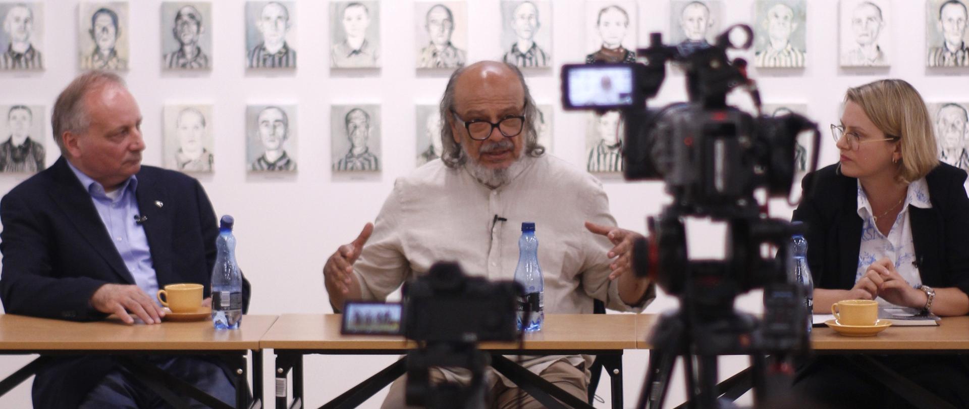 Fotografia przedstawia troje ludzi siedzących przy stole i rozmawiających przed kamerą telewizyjną. W tle jest wiele portretów z twarzami więźniów KL Auschwitz