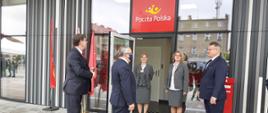 Na zdjęciu widać grupę ludzi stojących przed drzwiami punktu Poczty Polskiej. Wszyscy są uśmiechnięci. 