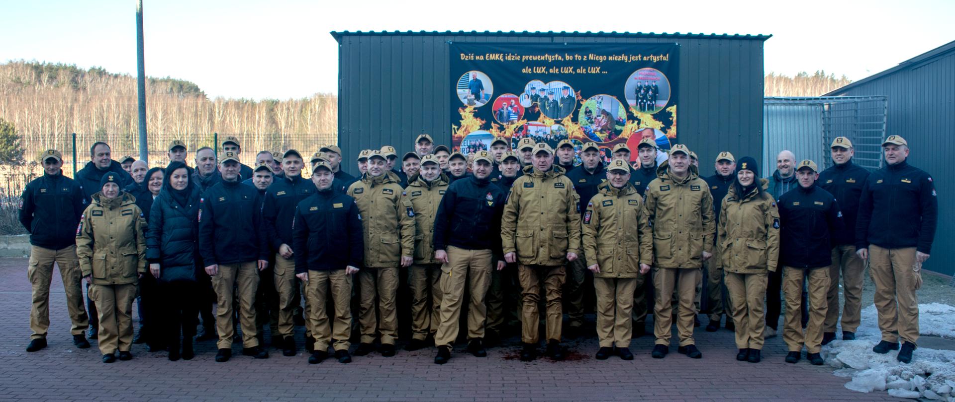 Przejście na zaopatrzenie emerytalne kapitana Aleksandra Grada. Funkcjonariusze Komendy Powiatowej Państwowej Straży Pożarnej w Brzezinach i zaproszeni goście uroczyście żegnają swojego kolegę, wspólnie pozują do zdjęcia, za pozującymi stoi wiata, na której zamieszczono nad ich głowami transparent ze zdjęciami zrobionymi podczas pełnionej służby kapitana Aleksandra Grada oraz napisem „Dziś na emkę idzie prewentysta, bo to z niego niezły jest artysta, ale lux, ale lux, ale lux”.