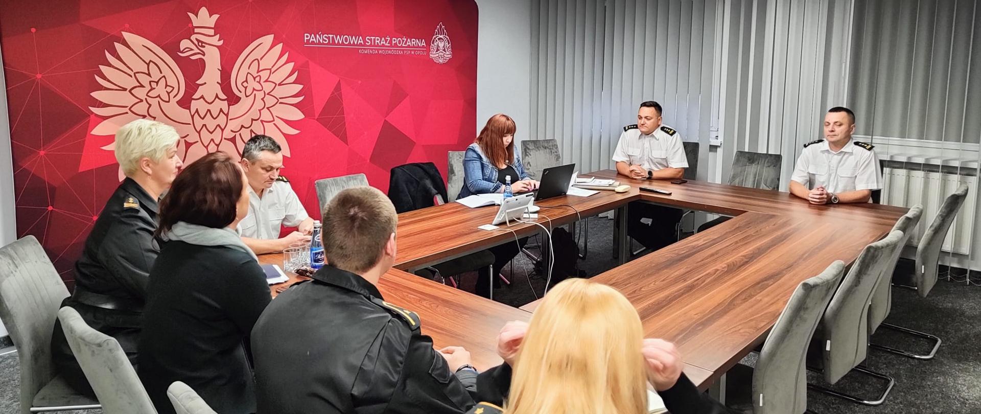 8, w tym 6 w umundurowaniu służbowym osób siedzących przy stołach w pomieszczeniu. W tle czerwony rollup z godłem oraz logiem Państwowej Straży Pożarnej.