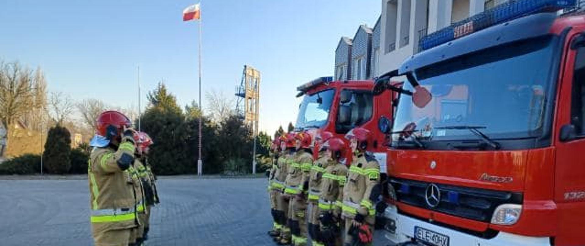 Na zdjęciu widać strażaków w żółtych strojach oraz czerwonych hełmach. Strażacy stoją w dwóch rzędach naprzeciwko siebie. Za jednym rzędem strażaków, za plecami, stoją dwa czerwone samochody strażackie. W tle widać masz, na którym jest flaga Polski, wspinalnie oraz krzewy. Z prawej strony widać kawałek budynku Komendy Powiatowej Państwowej Straży Pożarnej w Łęczycy. Jest widno.