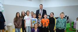 Minister Czarnek stoi w otoczeniu grupy kolorowo ubranych dzieci, za nimi biało-szara ściana.