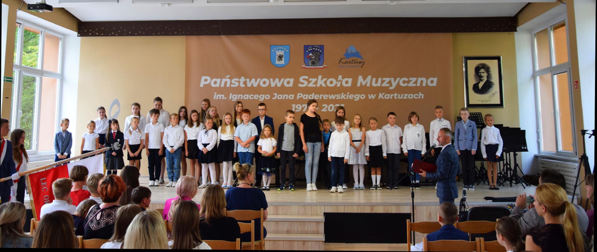 Na scenie znajdują się uczniowie, przed sceną poczet sztandarowy, dyrektor szkoły, rodzice i nauczyciele