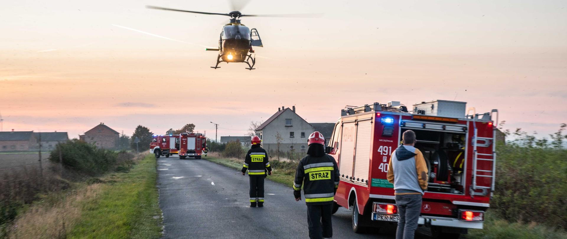 Nadlatuje śmigłowiec lotniczego pogotowia ratunkowego, który podchodzi do lądowania. Na jezdni trzy pojazdy pożarnicze z włączonymi sygnałami błyskowymi. Na jezdni stoi dwóch strażaków wskazując dogodne miejsce do lądowania. Osoba postronna przygląda się całej sytuacji. 