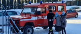 Na zdjęciu widać strażacki osobowy, terenowy samochód strażacki oraz trzy osoby w maseczkach: starszą kobietę podtrzymywaną za ręce przez strażaka ubranego w polar oraz przez kobietę w średnim wieku. Parking pokryty śniegiem.