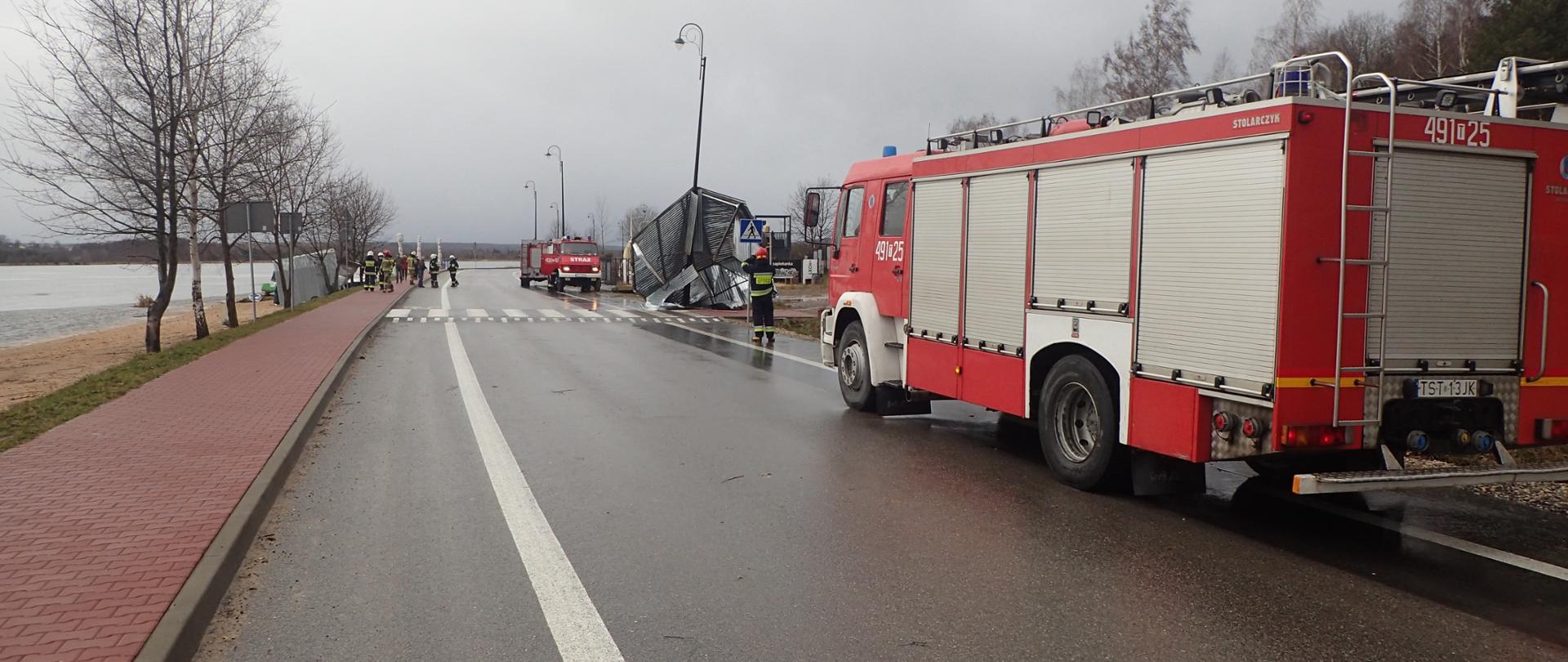 Blaszany garaż zabrany przez wiatr oparty o latarnię uliczną, przed garażem i w tle samochód strażacki, 