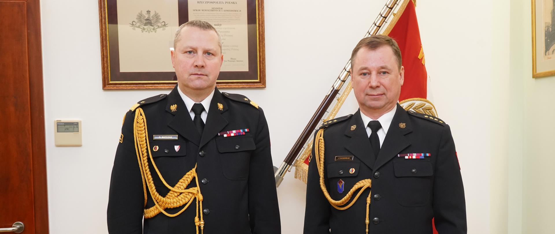 dwóch strażaków w ciemnych mundurach ze sznurem stoi obok siebie, strażak z prawej trzyma w ręku czerwoną teczkę, za nimi sztandar