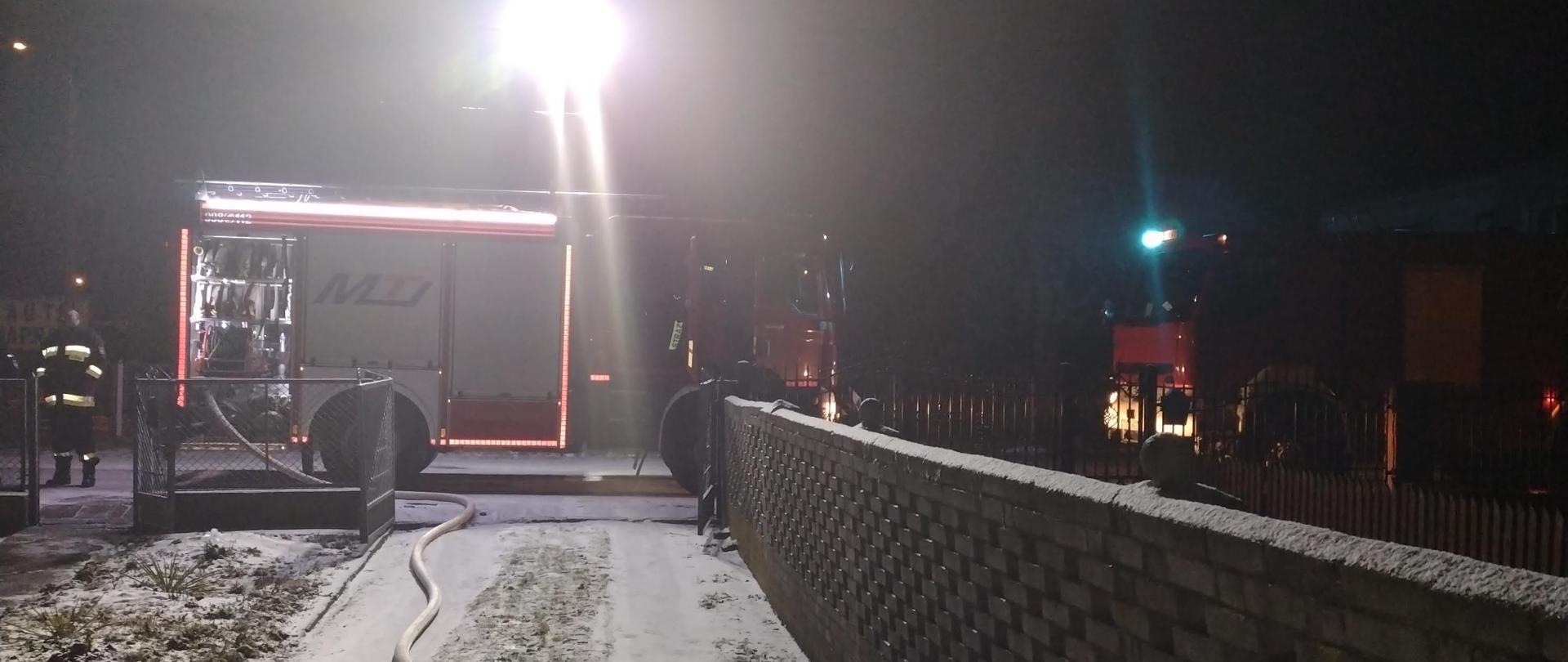 Na zdjęciu w porze nocnej widać zaparkowane samochody ratowniczo-gaśnicze przed budynkiem do którego doszło do pożaru. Samochody pożarnicze stoją przed wjazdem na posesję. Z jednego z samochodów rozwinięta jest linia gaśnicza.