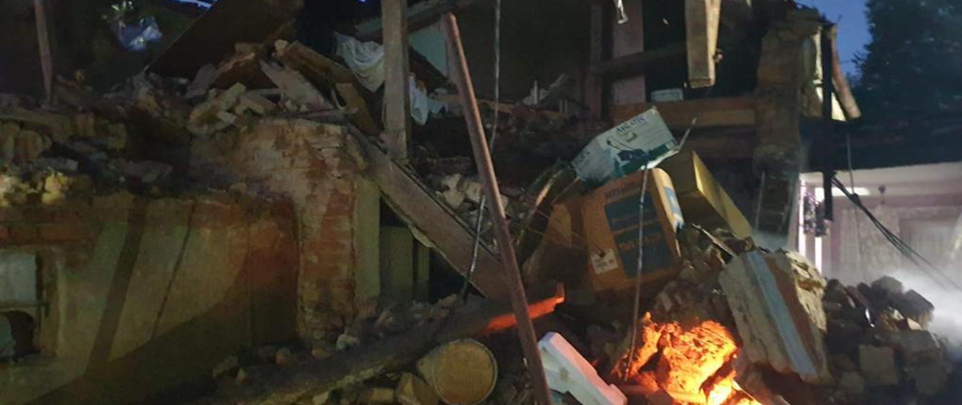 Zdjęcie wykonane porą nocną. Fotografia przedstawia gruz, oraz resztki konstrukcji budynku jednorodzinnego po wybuchu gazu. W lewym górnym rogu zdjęcia widoczny uszkodzony dach (liczne prześwity w konstrukcji dachu). W prawej części na gruzowisku widoczny niewielki płomień rozświetlający część gruzowiska.
