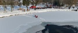 Dwóch strażaków na lodzie ciągnie specjalistyczne sanie ratunkowe w stronę przerębla, w którym znajduje się nurek. Akcja odbywa się w słoneczny, zimowy dzień na ośnieżonym akwenie. W tle widać gęsty tłum obserwujący ćwiczenia.
