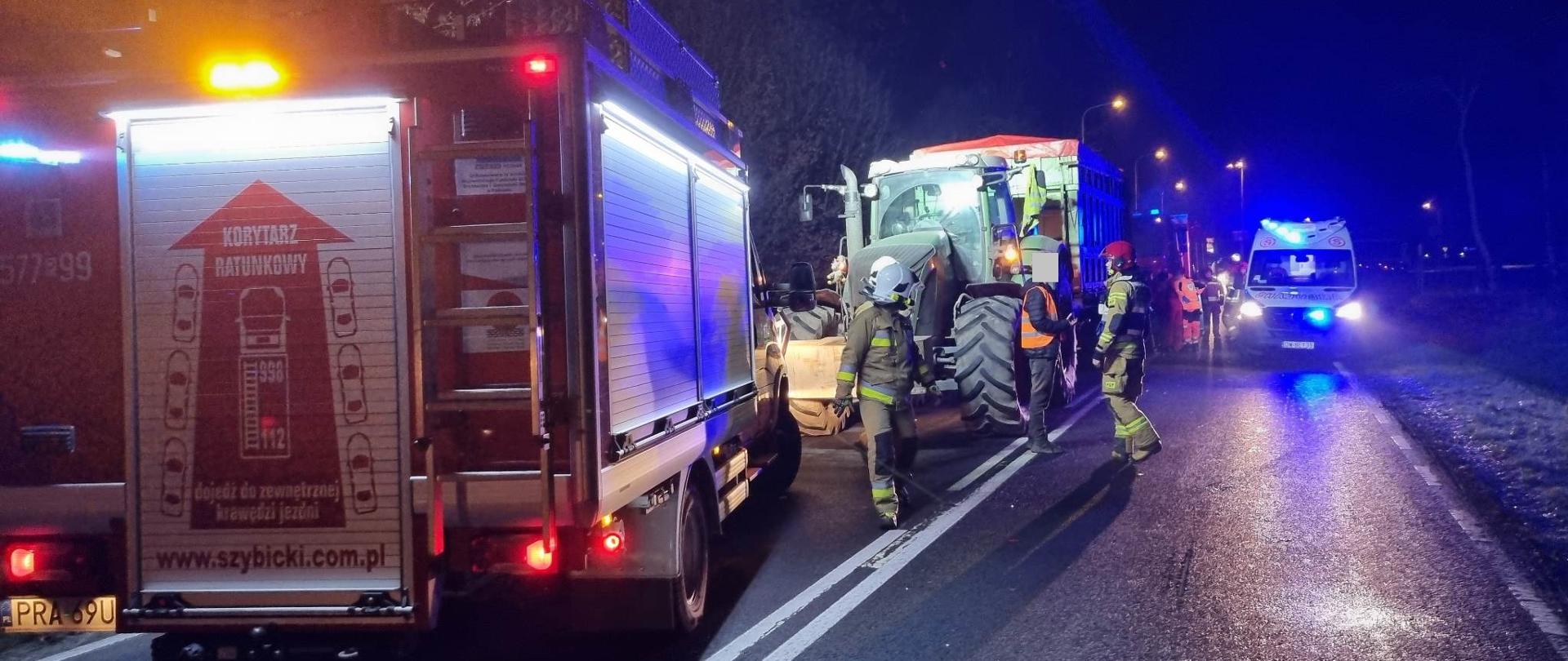Uderzenie samochodu osobowego w przyczepę rolniczą. Droga wojewódzka nr 309. Na pierwszym planie wóz straży pożarnej , który ma wysunięty i uruchomiony maszt oświetleniowy. W głębi ciągnik rolniczy z przyczepą, uczestniczące w wypadku. Z prawej strony ambulans medyczny. Wokół pojazdów przebywają strażacy i ratownicy medyczni. 