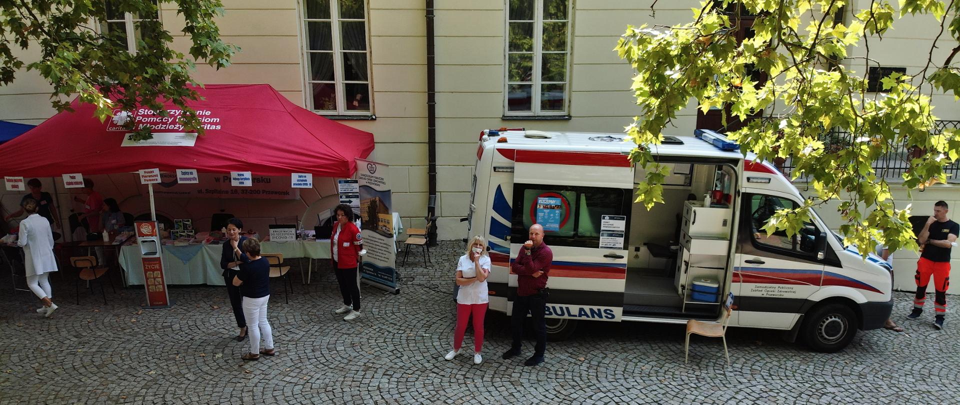 Na zdjęciu po prawej stronie znajduje się ambulans, w którym wykonywane są mobilne szczepienia, lewa strona zdjęcia ukazuje czerwony namiot, w którym prowadzone są zapisy. W kadrze ukazanych jest siedmiu sanitariuszy ubranych w uniformy medyczne, oraz jedna osoba cywilną. Ambulans jak i namiot znajdują się przed fasadą budynku.