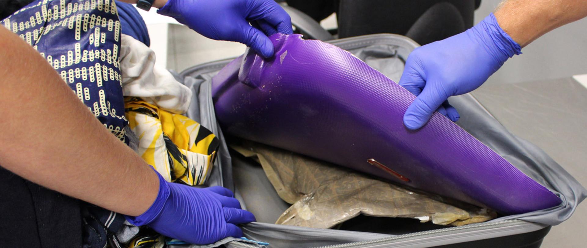 Dłonie w rękawiczkach rozchylają ścianki walizki gdzie ukryta jest paczka z heroiną.