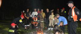 Obóz survivalowy w Sobiałkowie. Zdjęcie przedstawia rozpalone ognisko, wokół którego znajdują się uczestnicy obozu piekący kiełbaski. Sa to dziewczęta i chłopcy oraz osoby dorosłe. Wszyscy maja na sobie ubiory strażackie. Za ogniskiem stoi 5 meżczyzn, wśród których jest zastępca komendanta PSP, występujący w mundurze koloru piaskowego. 