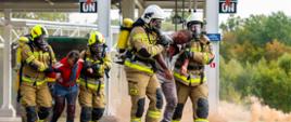 Dwie roty strażaków ewakuują osoby poszkodowane z widocznymi poparzeniami ze strefy niebezpiecznej. W tle, za nimi widoczny pojazd cysterny i kłęby ciemnego dymu.