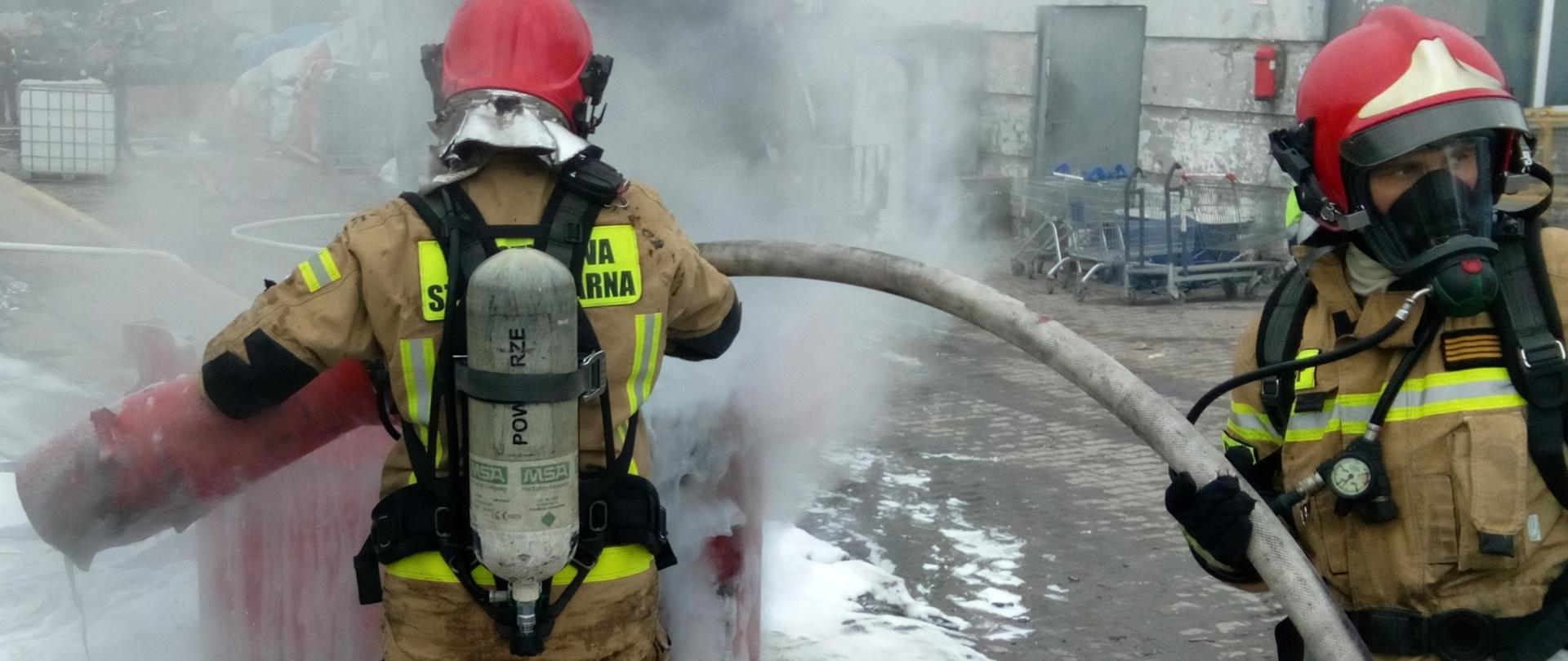 Zdjęcie przedstawia dwóch strażaków podających pianę na metalowy pojemnik, w którym palą się baterie.