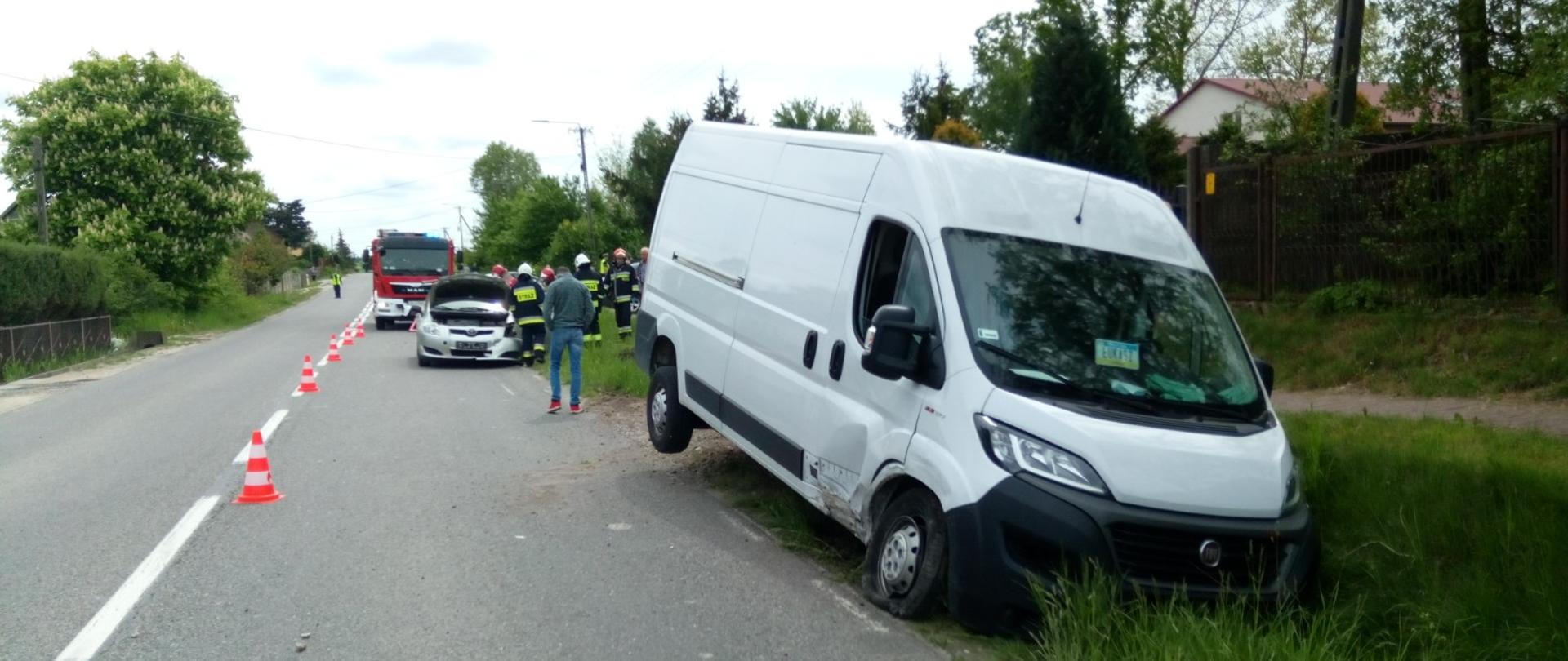 Zdjęcie przedstawia drogę na której doszło do zderzenia dwóch pojazdów. Dostawczy lekko uszkodzony biały bus znajduje się poza pasem jezdni w przydrożnym rowie. W oddali widać samochód strażacki oraz drugi z samochodów z podniesioną pokrywą silnika biorący udział w zdarzeniu. Wokół pojazdów znajdują się strażacy.