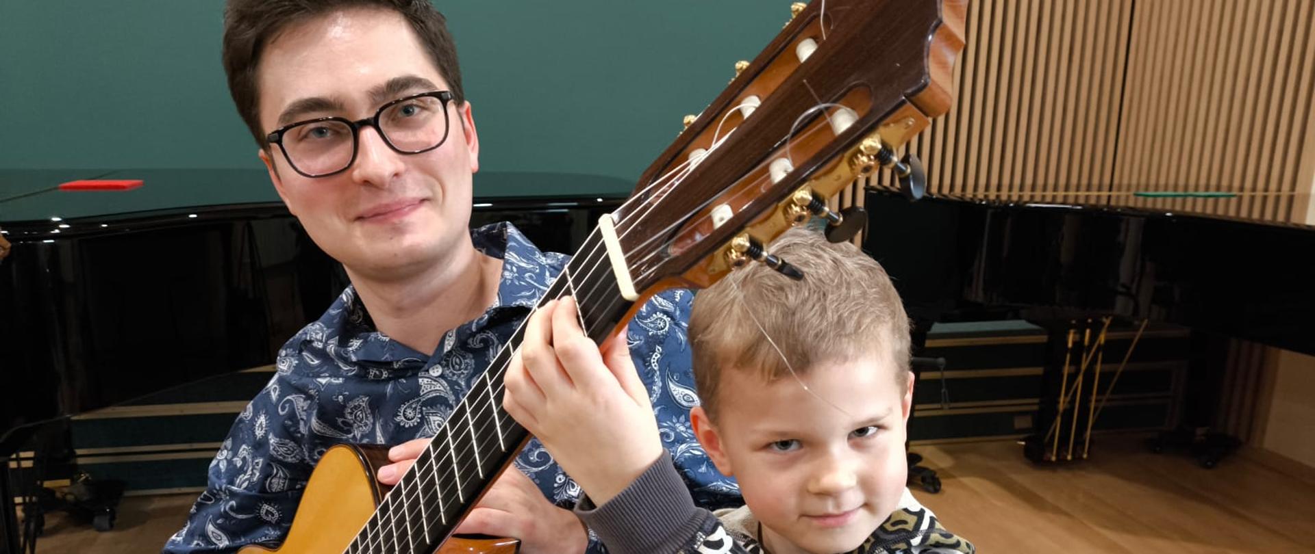 Na zdjęciu widać gitarzystę który trzyma gitarę w prawej ręce a obok niego stoi uczeń który trzyma gryf lewą ręką za nimi stoi fortepian oraz jest widoczna Zielona ściana z drewnianymi lamelami. Na podłodze po prawej stronie widoczny fragment czerwonego futerału na gitarę