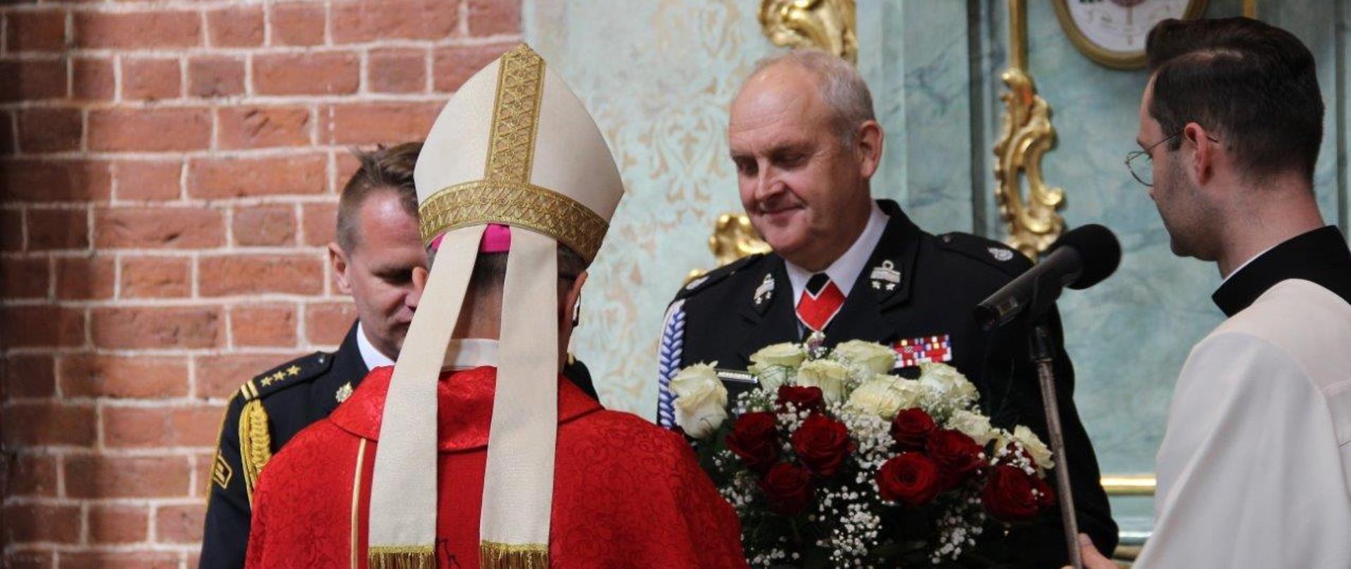 Oficer PSP i strażak OSP w mundurach galowych, stoją przed biskupem i wręczają upominki w podziękowaniu za eucharystię. Strażak z prawej strony trzyma kwiaty biało-czerwone, strażak z lewej statuetkę świętego Floroana.