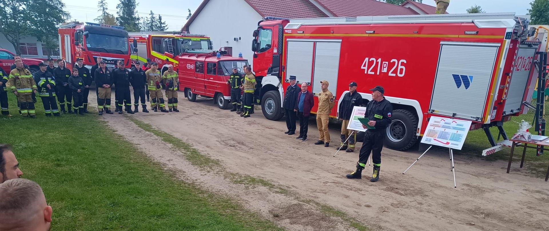 Na zdjęciu widać rozpoczęcie warsztatów szkoleniowych Integracja 2024. Strażacy ustawieni są w kształcie litery U, witani są przybyli goście natomiast w tle widać samochody pożarnicze