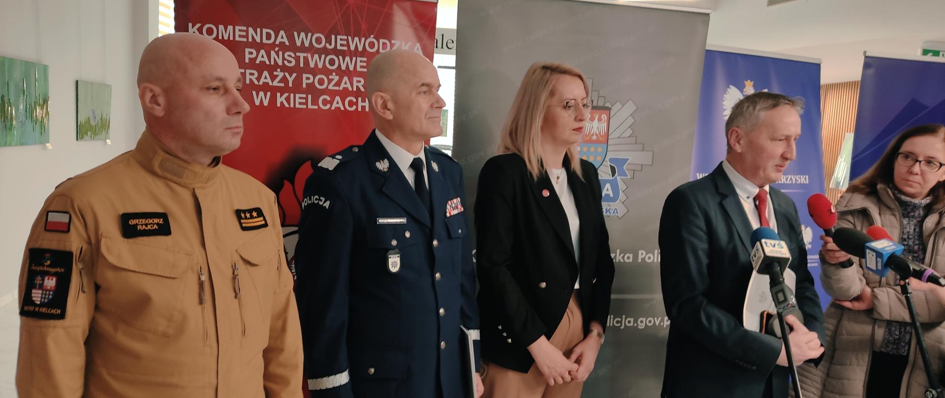 Na zdjęciu widzimy 4 osoby stojące na ściankach podczas konferencji prasowej. Pierwszy o lewej strażak w piaskowym ubraniu, potem generał policji, kobieta w stroju wizytowym oraz mężczyzna w garniturze.