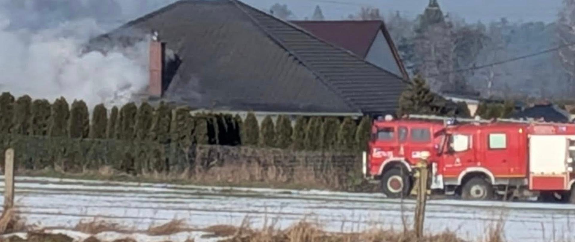 Na zdjęciu znajduje się dom jednorodzinny z którego dachu unosi się dym oraz samochód pożarniczy