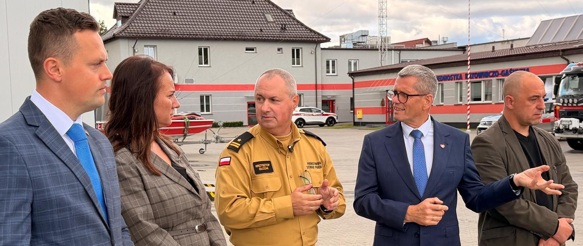 Na terenie strażnicy pożarnej pięć osób stoi na placu przed budynkami remizy. Wśród nich znajduje się strażak w mundurze oraz cztery osoby w ubraniach cywilnych. W tle widać budynki, maszt telekomunikacyjny, drzewa oraz niebo z dużymi chmurami i przejaśnieniami.