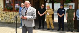 Przed mikrofonem stoi mężczyzna ubrany w garnitur, za nim przedstawiciele rożnych służb. Z tyłu żółty budynek z wiatą.