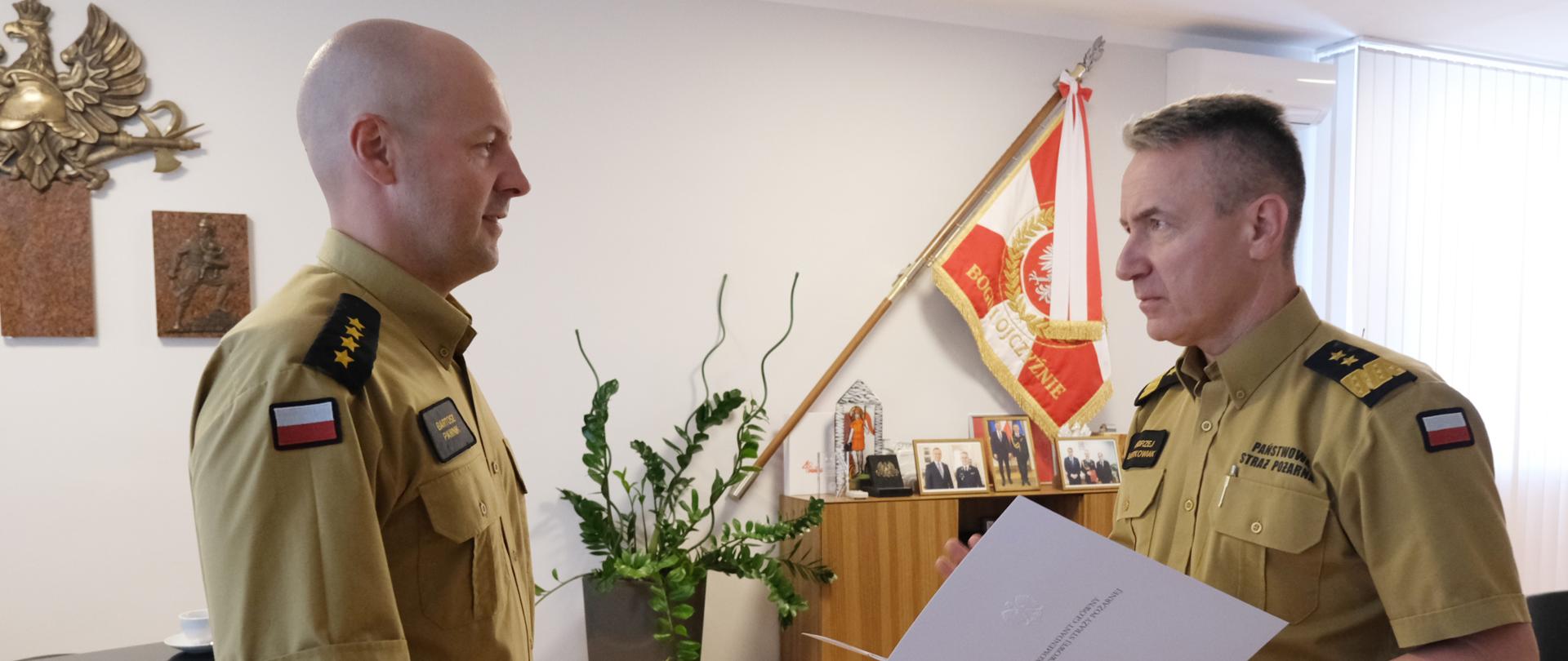 Dwaj mężczyźni w piaskowych mundurach. Stojący po prawej trzyma w dłoniach białą teczkę