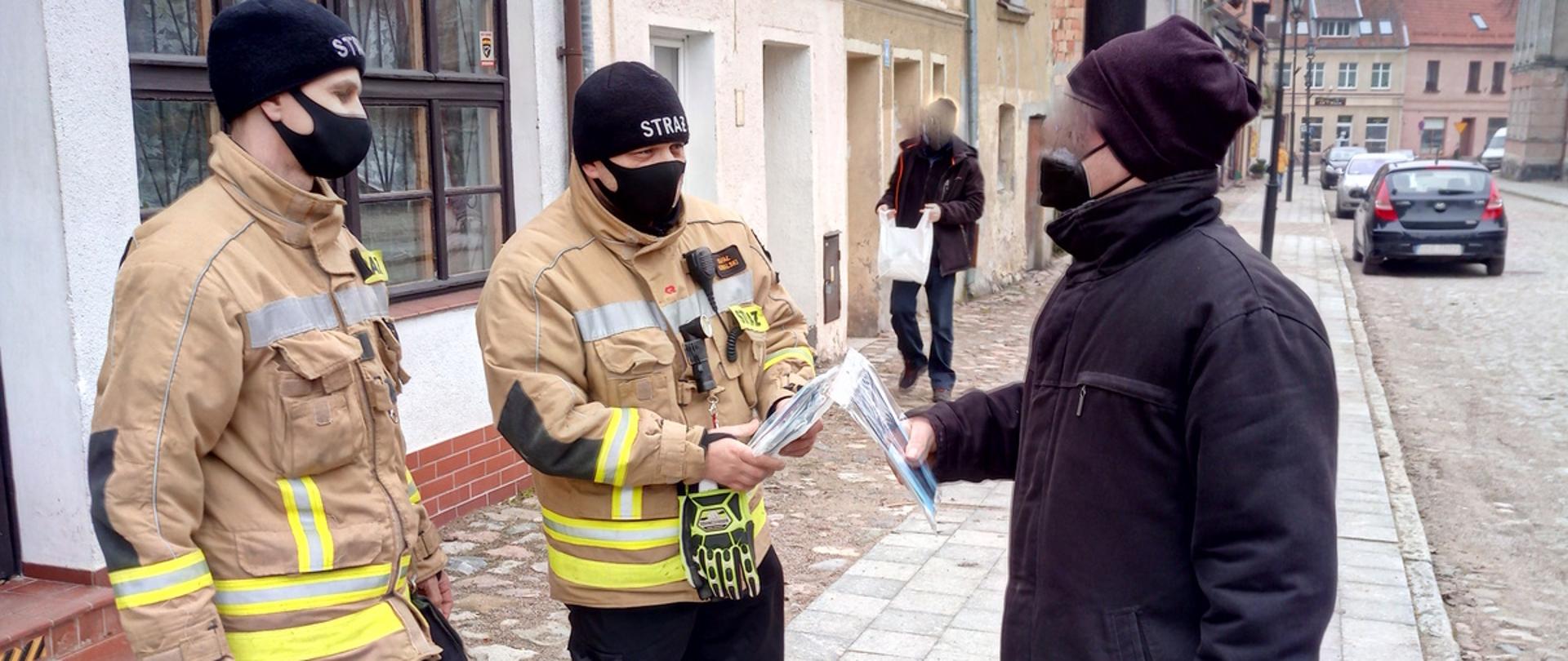 Zdjęcie przedstawia dwóch strażaków wręczających pakiet (maseczka + ulotka) mieszkańcowi Reszla. Strażacy w czarnych ubraniach koszarowych, czarnej czapce, maseczce oraz piaskowej bluzie ubrania specjalnego. Mieszkaniec w czarnej kurtce, spodniach jeansowych trzyma w jednej ręce białą reklamówkę a drugą ręką odbiera pakiet. Osoby stoją na chodniku a w tle widać stare kamienice. 