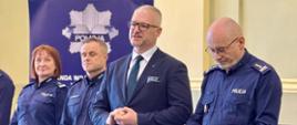 Mężczyzna w garniturze, wojewoda, przemawia na tle ścianki Komendy policji 