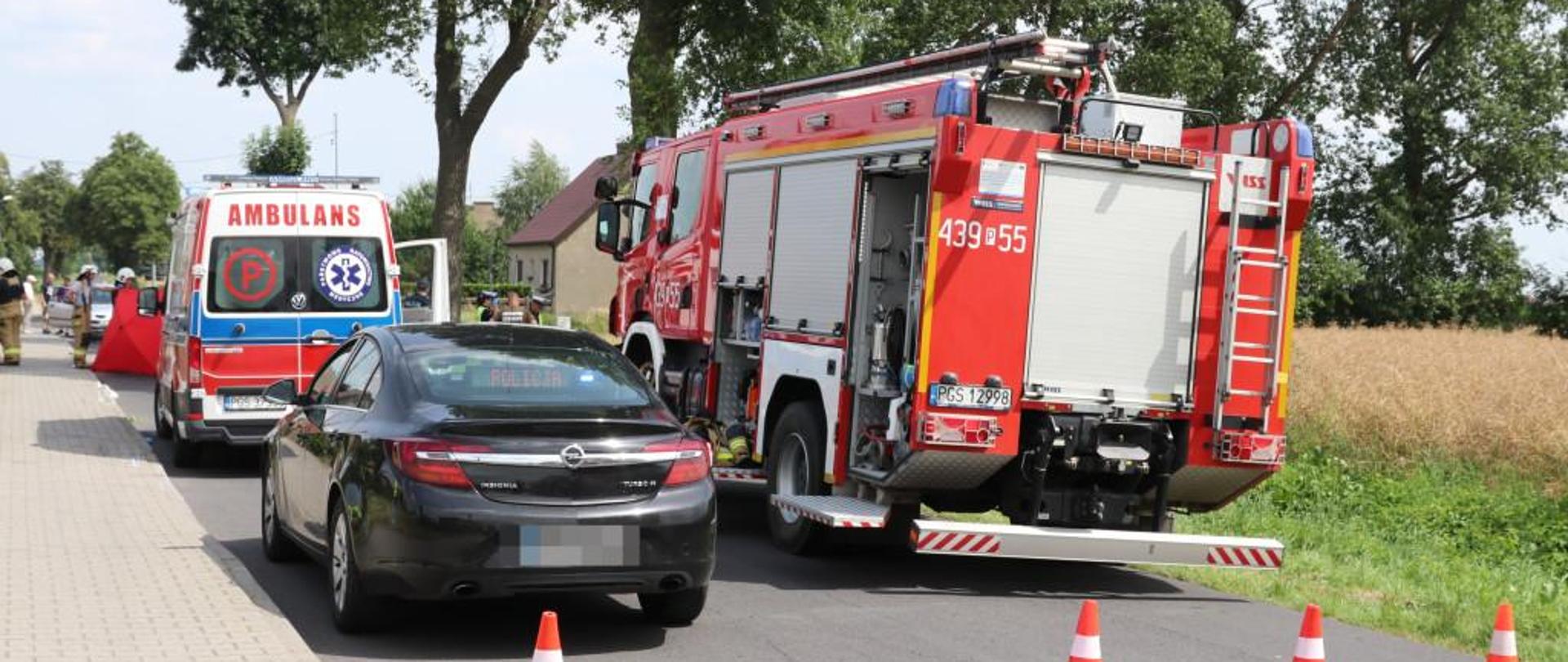 Wóz strażacki, karetkę pogotowia oraz nieoznakowany samochód policji. Pojazdy stoją na drodze podczas zdarzenia. Przed nimi stoją 4 pachołki. W tle stoją strażacy, którzy trzymają parawan.