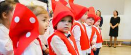 Pod ścianą stoi grupa małych dzieci w czerwonych kubraczkach i czapeczkach.