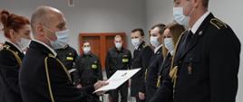 Komendant powiatowy mł. bryg. Arkadiusz Makowski ubrany w mundur galowy ze sznurem w maseczce ochronnej na twarzy wręcza awanse strażakom tutejszej komendy, w lewej dłoni trzyma teczkę na akta a prawą dłoń trzyma w uścisku strażaka, w głębi funkcjonariusze stoją w szeregu, na wprost trzech strażaków w pozycji zasadniczej ubranych w ubranie koszarowe w maseczkach ochronnych na twarzy