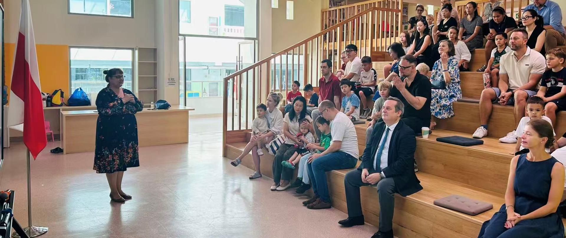 Polska szkoła w Szanghaju, rozpoczęcie roku szkolnego, po lewej przy fladze, stoi dyrektorka szkoły, po prawej na wysokich stopniach siedzi ok. 30 osób - dzieci i rodzice. 
