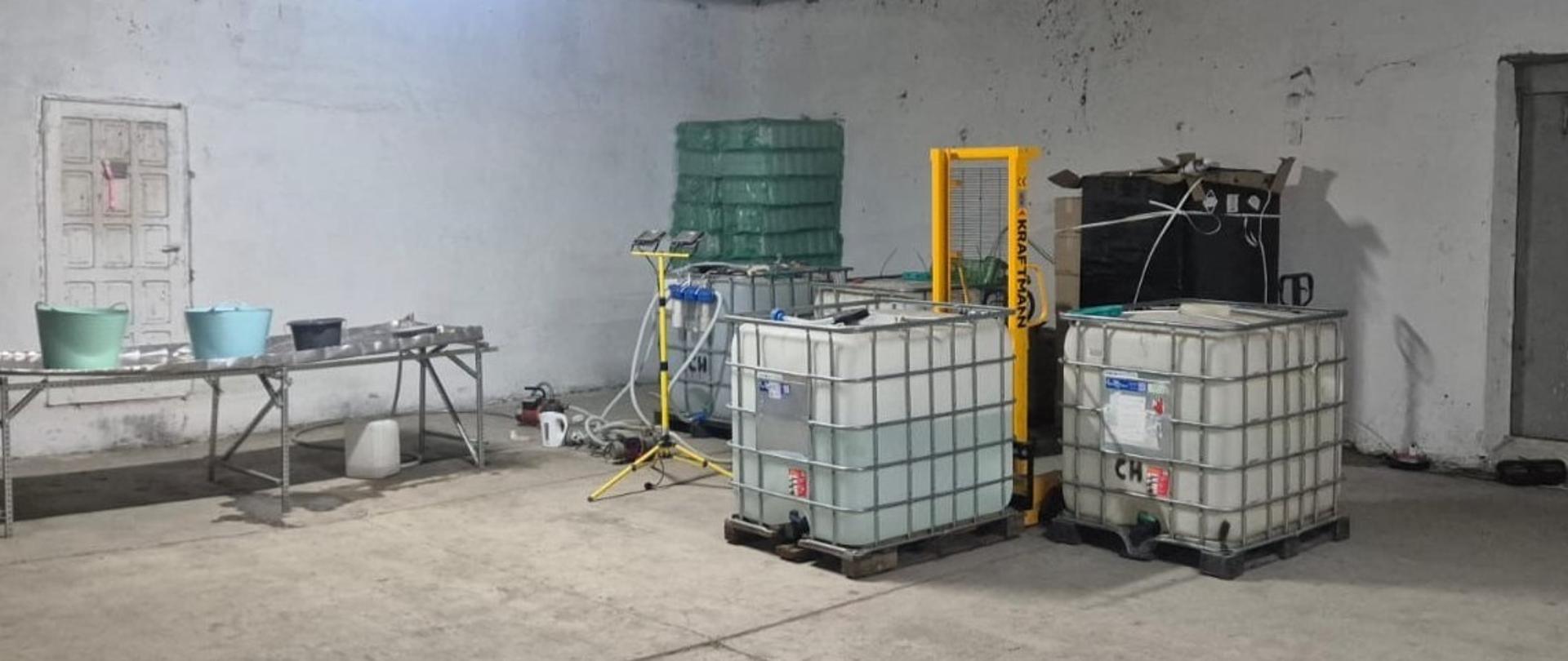 W magazynie znajdują się dwa duże zbiorniki, stół roboczy oraz urządzenia techniczne. Obok zbiorników stoi ręczny podnośnik paletowy.