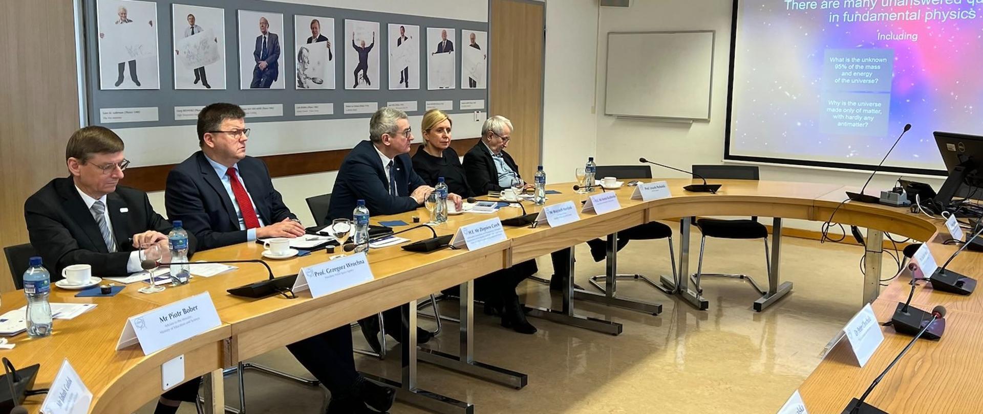 Przy dużym owalnym stole siedzi kilka osób, patrzą na duży monitor wiszący na ścianie nad stołem.